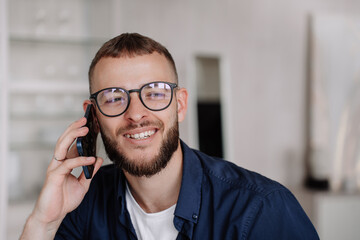 Close up of young beardy American man talks by phone looks aside smiles. Cheerful caucasian guy in glasses holds smartphone at home. Successful business man speaks with partner. Handsome male at hotel