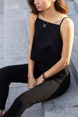 Young stylish happy brunette woman sitting on a busy business urban street in black sleeveless top and pants. Street style photoshoot.
