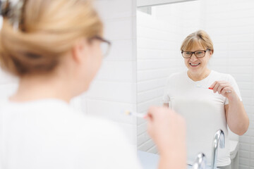 Obraz premium Happy caucasian woman brushing teeth and looking at mirror in bathroom