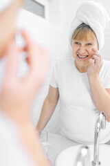 Fototapeta premium Gorgeous mid age older adult 50 years old blonde woman wears bathrobe in bathroom applying nourishing antiage face skin care cream treatment, looking at mirror doing daily morning beauty routine.