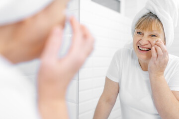 Fototapeta premium Gorgeous mid age older adult 50 years old blonde woman wears bathrobe in bathroom applying nourishing antiage face skin care cream treatment, looking at mirror doing daily morning beauty routine.