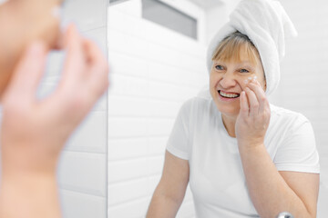 Gorgeous mid age older adult 50 years old blonde woman wears bathrobe in bathroom applying nourishing antiage face skin care cream treatment, looking at mirror doing daily morning beauty routine.