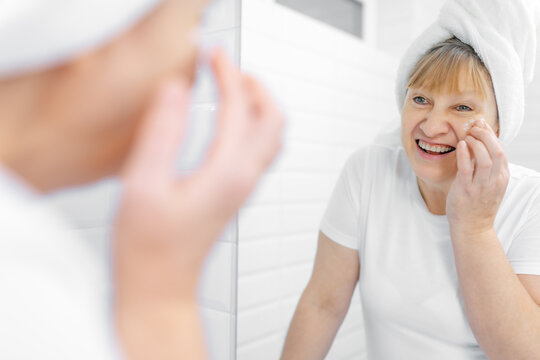 Gorgeous Mid Age Older Adult 50 Years Old Blonde Woman Wears Bathrobe In Bathroom Applying Nourishing Antiage Face Skin Care Cream Treatment, Looking At Mirror Doing Daily Morning Beauty Routine.