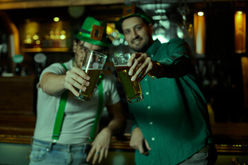 Two friends toasting with beers in a bar to celebrate St. Patrick's Day. Green lighting throughout the bar.