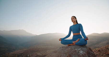 Naklejka na ściany i meble Asian woman wearing active wear do yoga practice meditating in mountains, lotus position. No stress, mindfulness, inner balance concept.Woman Yoga - relax in nature. Copy space.