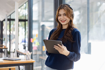 Fototapeta premium Young woman smiling and holding digital tablet with earphones standing in office, People And Technology Concepts. 