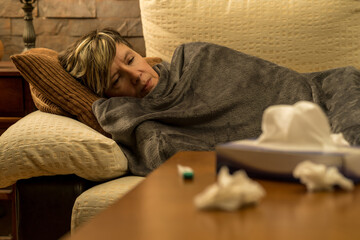 mujer mediana edad enferma con dolores  en el sofa de casa tapada con una manta, tomando medicina...