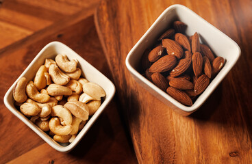 Two white ceramic bowls with cashew and almonds nuts on a wooden table.