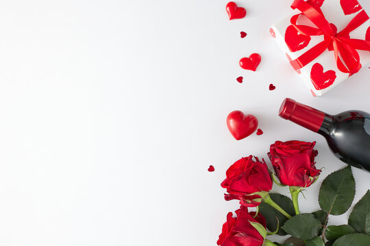 Valentine's Day presents concept. Top view photo of red roses, wine bottle, gift box and red hearts on white background with copy space. Holiday card idea.