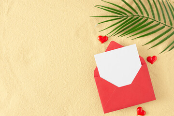 Summer holiday concept. Flat lay photo of open envelope with white card, tropical leaf, red hearts on sandy background with empty space. Travel holiday card idea.