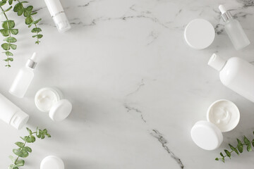Skin care beauty concept. Flat lay photo of white cosmetic tubes, cream jars, dropper bottles and eucalyptus leaves on marble background with empty space in the middle. Cosmetics mockup idea.