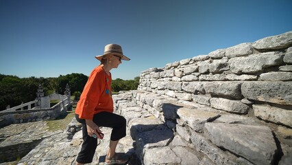 Mature woman wearing ethnic clothes, sunglasses, hat holding phone climbing stone stairs to the top of Xcambo Mayan pyramid.