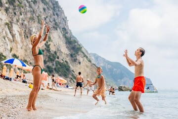 Mom in a green bathing suit and a straw sun hat plays ball with her sons on the beach. Dream vacation in Corfu, Greece
