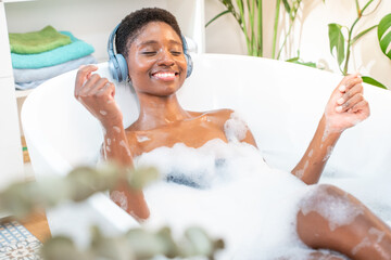 Attractive black woman wearing headphones happy relaxing and dancing in foam bath in beautiful...