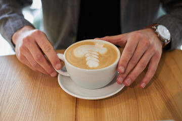 Close up of  white ceramic cup of coffee latte or cappuccino with a decoration on the top of foam, in the hands of a businessman entrepreneur while a coffee break. Hot drink concept 