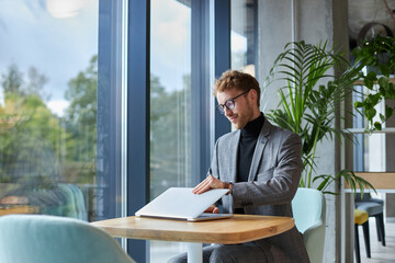 Handsome young Caucasian businessman in formal suit, turning on his laptop in a light modern office