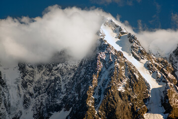 White clouds cover the tops of high rocky mountains with snow and glaciers in Altai in the autumn afternoon.