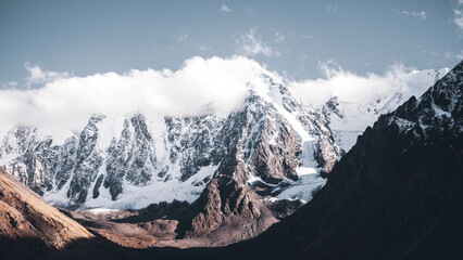 White clouds cover the tops of high rocky mountains with snow and glaciers in Altai with fog.