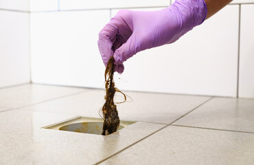 Water drain hole is clogged with hair clump. Woman hand in gloves is cleaning the bathroom sewer...