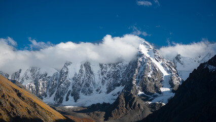 Fototapeta premium White clouds cover the tops of the mountains with snow and glaciers in Altai in the shadow of the gorge with fog.