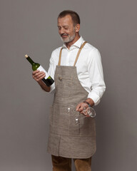 Respectable man in a linen apron holds wine glasses and a bottle of wine. Sommelier or waiter in joy.