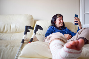 Young woman on couch at home with crutches in orthopedic plaster videochating on the phone. Fracture of broken leg foot or knee. Concept of rehabilitation and healing. Orthopedics and Traumatology. - obrazy, fototapety, plakaty