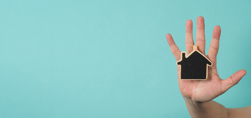 Small wood house in hands represent concepts such as home care family love real estate housing shelter insurance and mortgage. Hands holding small model house isolated on blue green studio background.