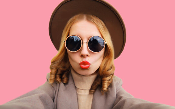 Fashionable portrait of stylish young woman stretching hand for taking selfie with smartphone blowing her lips with red lipstick sending sweet air kiss on pink background