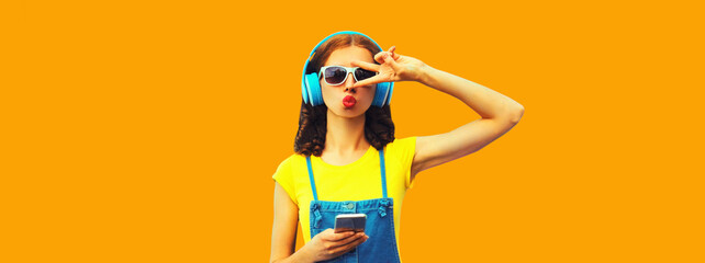 Portrait of stylish young woman in headphones listening to music with smartphone on yellow background
