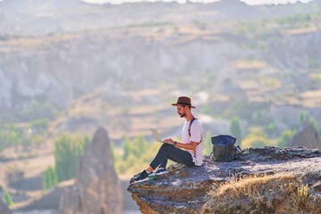 Man traveler blogger works remotely at computer sitting on a cliff at calm destination. Work...
