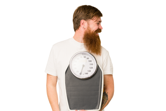 Adult redhead man with long beard holding a scale isolated cut out looks aside smiling, cheerful and pleasant.