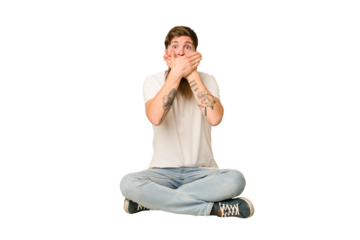 Young adult redhead with a long beard sitting on the floor isolated shocked covering mouth with hands.