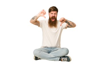 Young adult redhead with a long beard sitting on the floor isolated showing thumb down and expressing dislike.