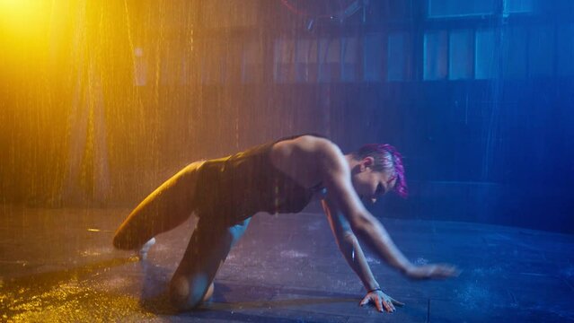 Sexy seductive slim woman dancing under pouring rain, moving passionately in darkness. Go-go dance performance and peep show in nightclub by energetic music in futuristic yellow blue glowing spotlight