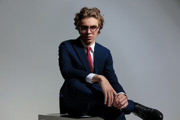 curly hair man in elegant suit holding leg over knee in front of grey background