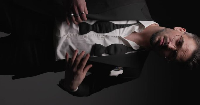 sexy young groom with undone bowtie buttoning tuxedo, smiling and looking to side while posing on grey background