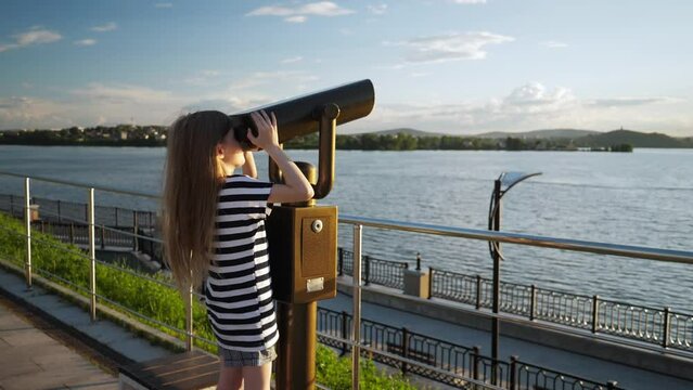 Slow-motion footage of little blonde girl looking at sights of opposite shore through fixed city binoculars while standing on observation deck