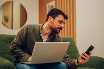 Caucasian bearded man using laptop and a smartphone at home on a couch