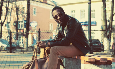 Portrait of stylish young african man model posing wearing black rock leather jacket with bag sitting on the bench on city street background