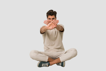 Young caucasian man sitting on the floor isolated on white background doing a denial gesture