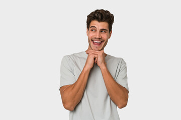 Young handsome caucasian man isolated on white background keeps hands under chin, is looking happily aside.