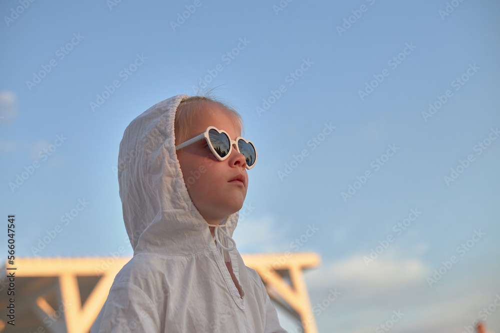 Wall mural young child with glasses in a white hood against the sky. Portrait of a young girl at sunset in the evening