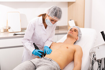 Obraz na płótnie Canvas Confident female beautician performing body ultrasound cavitation procedure for young man at aesthetic clinic