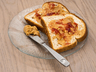 Two classical American sandwiches with peanut butter and jelly on a glass plate with a knife with peanut butter on wooden board.