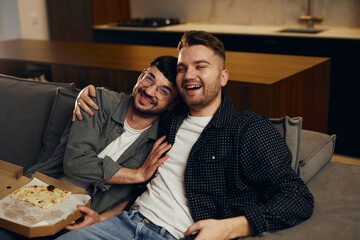 Two gay men in casual clothes are sitting on the sofa, hugging and watching TV at home. High quality photo