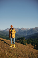 Woman autumn full-length walk on the hill smile with teeth and look at the mountains in a yellow raincoat and jeans happy hiking trip, freedom lifestyle 