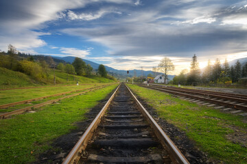 Fototapeta na wymiar Railroad in mountains at sunset in summer. Beautiful industrial landscape with railway station, trees, green grass, blue sky, clouds in spring. Old rural railway platform in Ukraine. Transportation