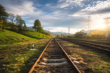 Naklejka na ściany i meble Railroad in mountains at sunset in summer. Beautiful industrial landscape with railway station, trees, green grass, blue sky, clouds in spring. Old rural railway platform in Ukraine. Transportation