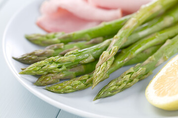 Fresh cooked green asparagus with ham slices and lemon on blue plate (Selective Focus, Focus on parts of the five asparagus heads in the front)