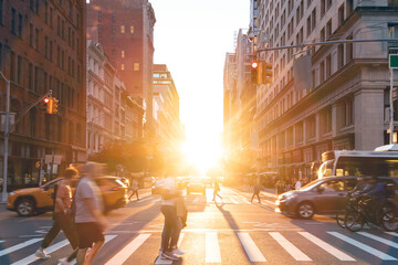 People, cars, bicycles and buses traveling through a busy intersection on 5th Avenue and 23rd Street in New York City with sunlight shining between background buildings